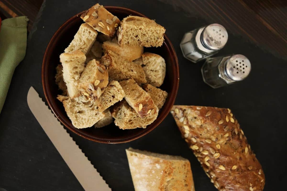 Cubed bread on a plate with cut loaves, a bread knife, and a salt and pepper shaker surrounding it.