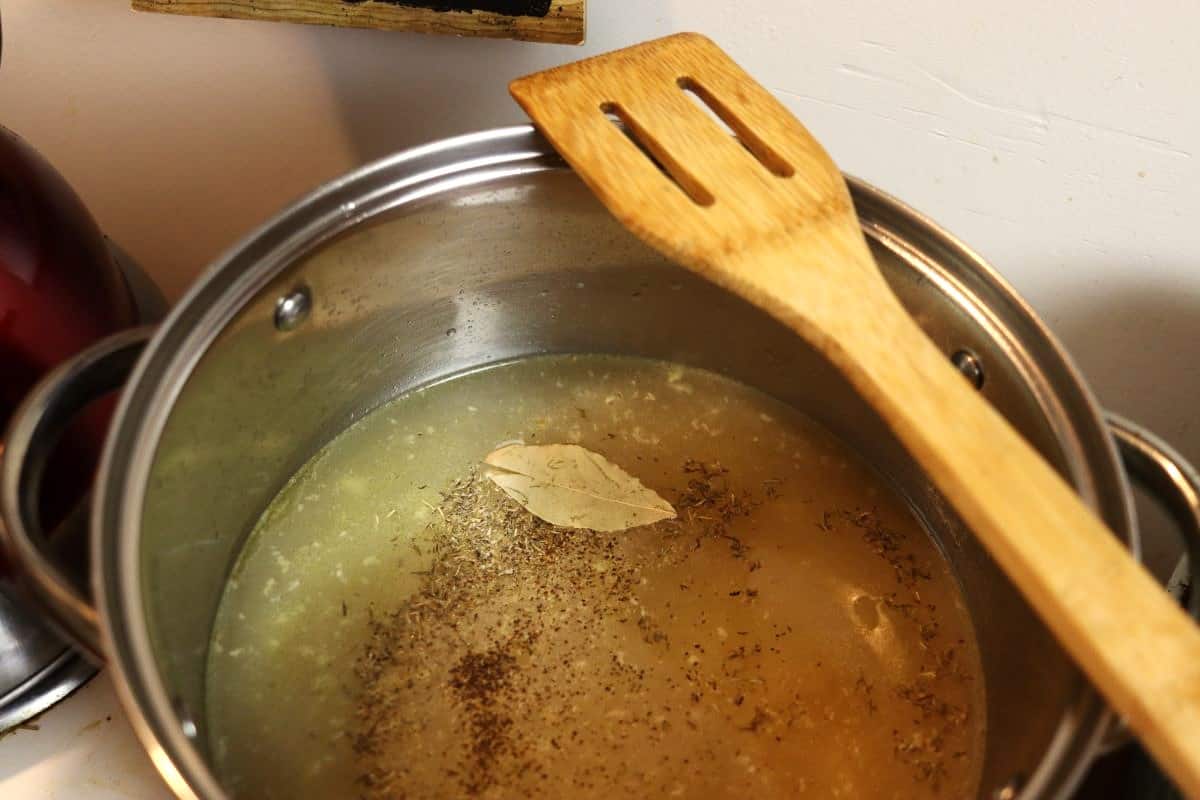 Pot full of golden broth with seasoning and a bay leaf floating on top and a woooden spoon sitting on the edge of the pot.