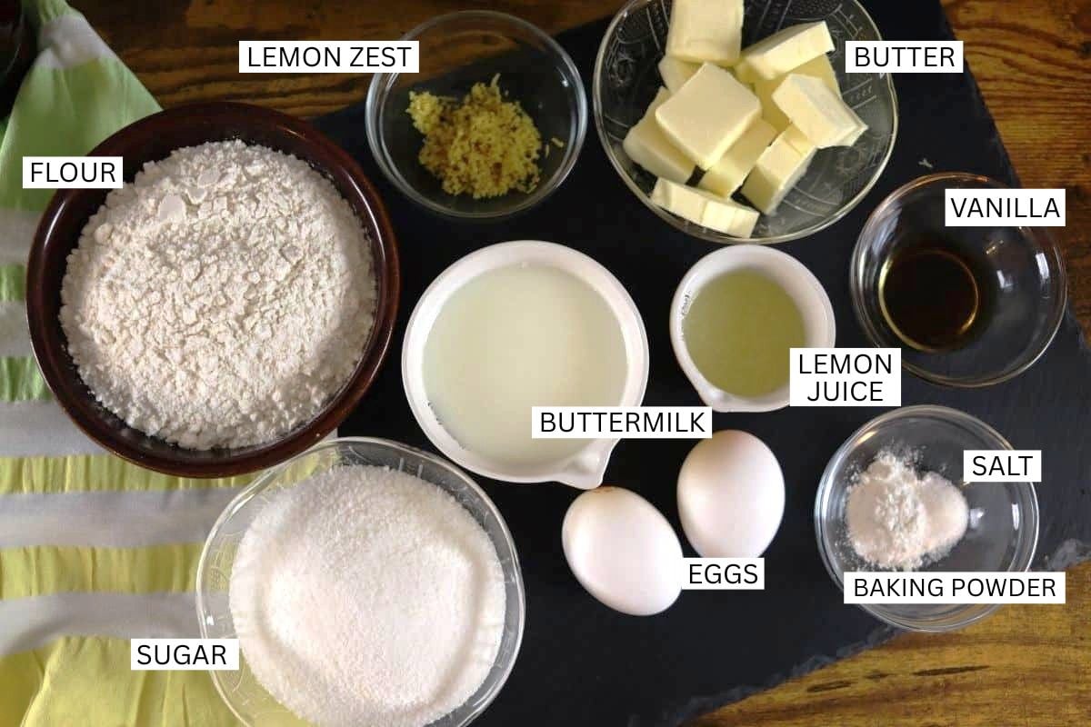 Bowls labeled with ingredients for baking, including flour, sugar, lemon zest, butter, vanilla, buttermilk, lemon juice, eggs, salt, and baking powder, arranged on a dark surface.