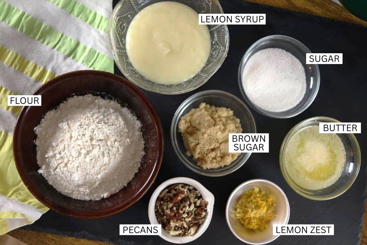 Small bowls holding labeled baking ingredients: lemon syrup, sugar, brown sugar, melted butter, flour, chopped pecans, and lemon zest, arranged on a dark surface with a striped towel.