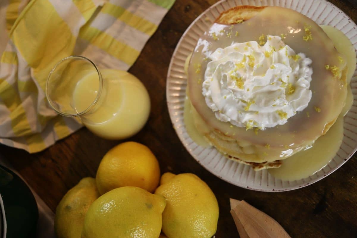 A plate of pancakes topped with lemon sauce, whipped cream, and lemon zest sits on a wooden table beside fresh lemons, a glass of lemon curd, a striped cloth, and a wooden citrus reamer.