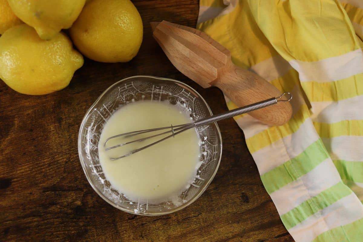 a bowl of lemon syrup and cornstarch with a whisk in it sitting on a countertop next to a striped cloth, fresh lemons, and a wooden handheld juicer.