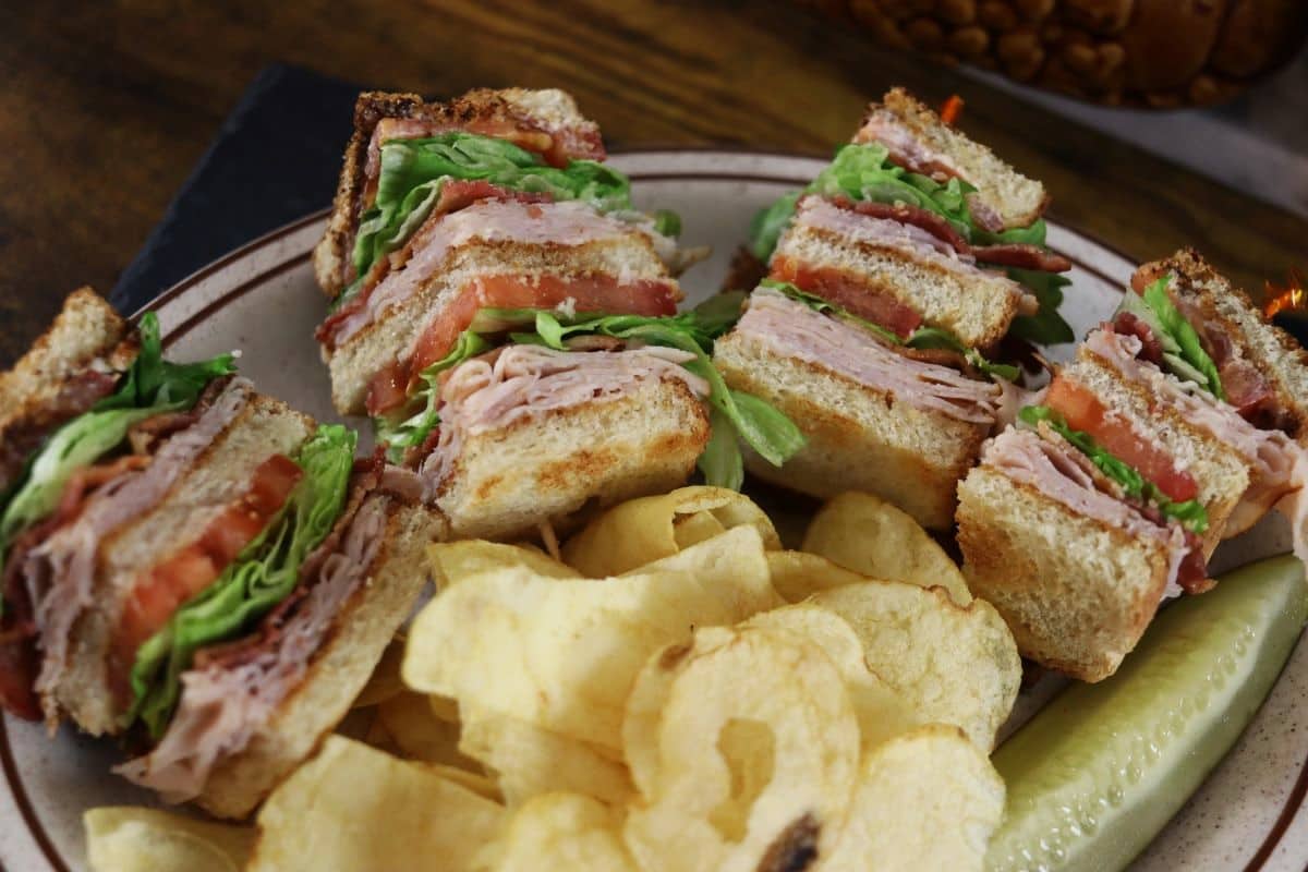 A plate with four stacked club sandwich quarters filled with turkey, lettuce, tomato, and bacon, served with potato chips and a dill pickle spear.