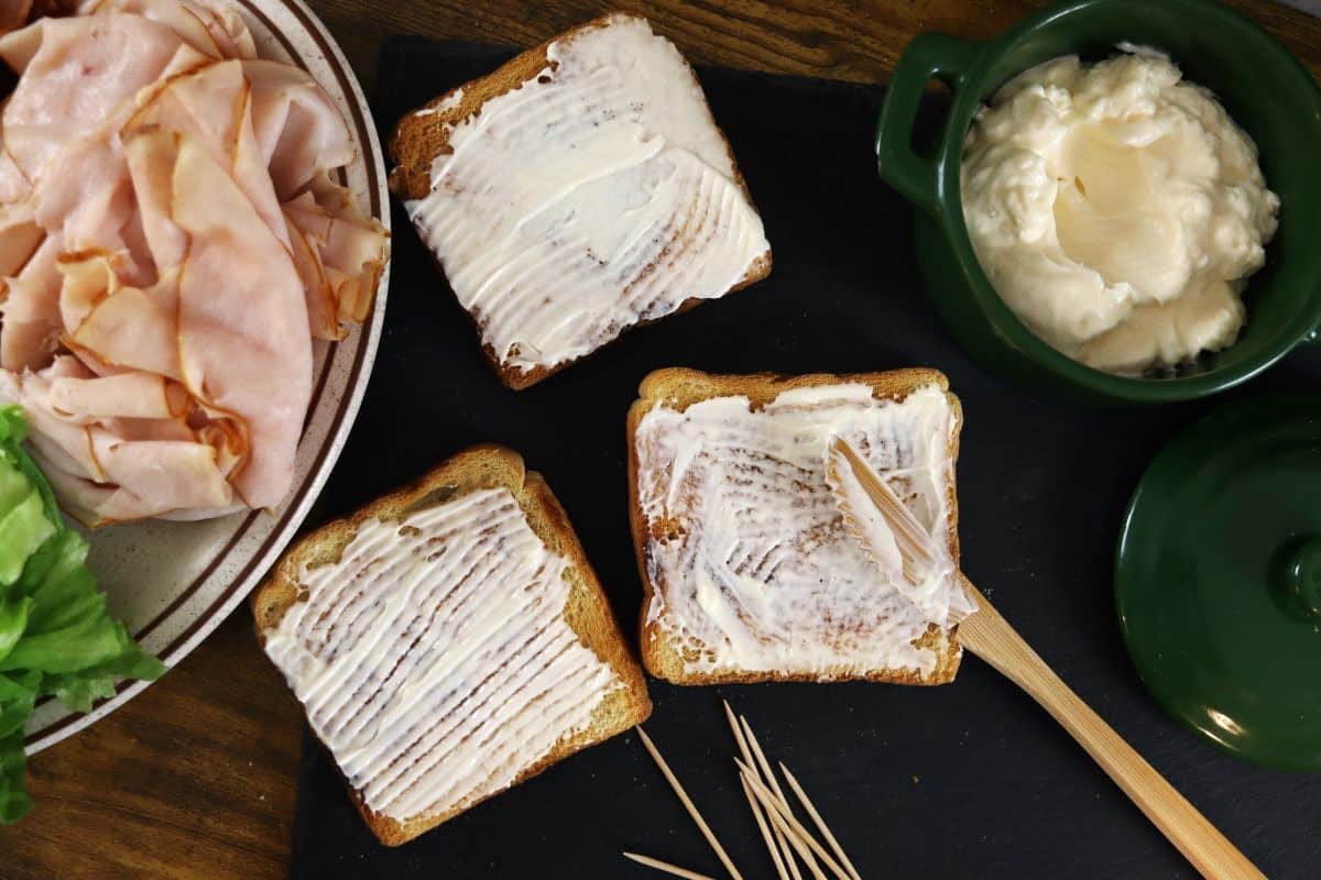 3 slices of square toast on a slate board with mayonnaise spread on them sitting next to a dish of mayo and a plate of turkey and with a wooden knife.