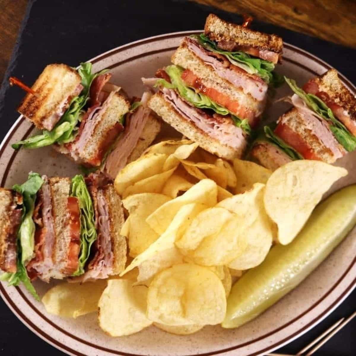 Four stacked club sandwich quarters filled with turkey, lettuce, tomato, and bacon, on a plate with potato chips and a dill pickle spear.