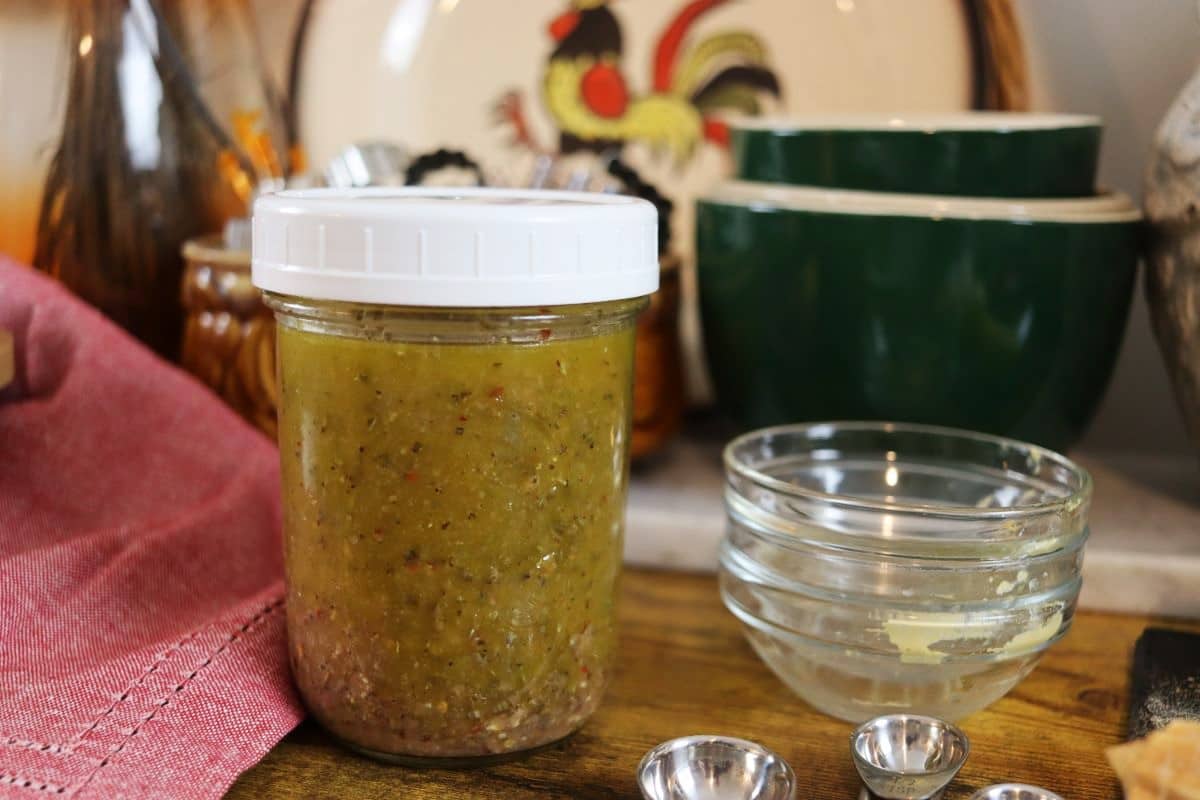mason jar of golden colored dressing with herbs init on a table next to glass condiment dishes.
