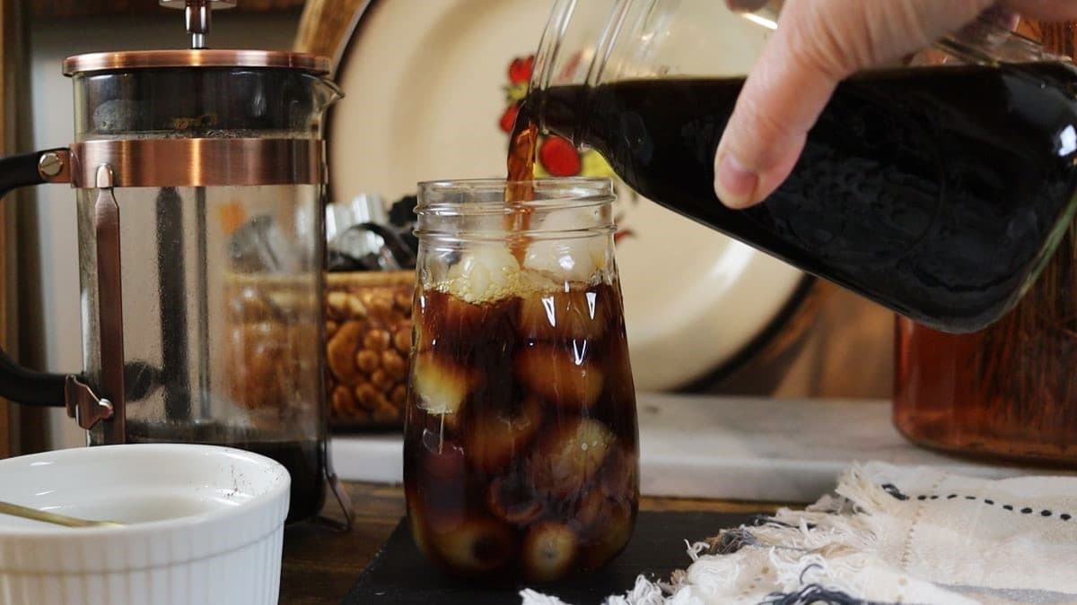 A hand pours dark cold brew coffee from a glass carafe into a mason jar filled with ice. A French press, a bowl, and a cloth napkin are on the wooden table nearby.