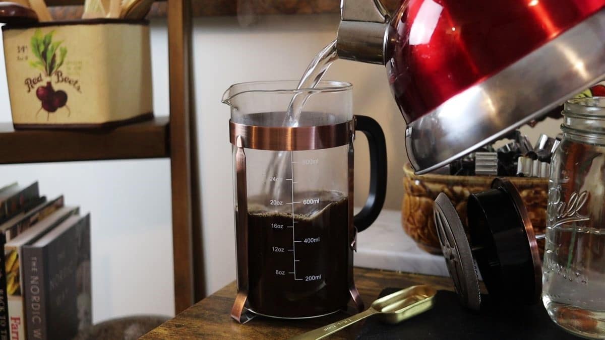 pouring hot water from a teapot into a french press coffee maker full of coffee grounds.