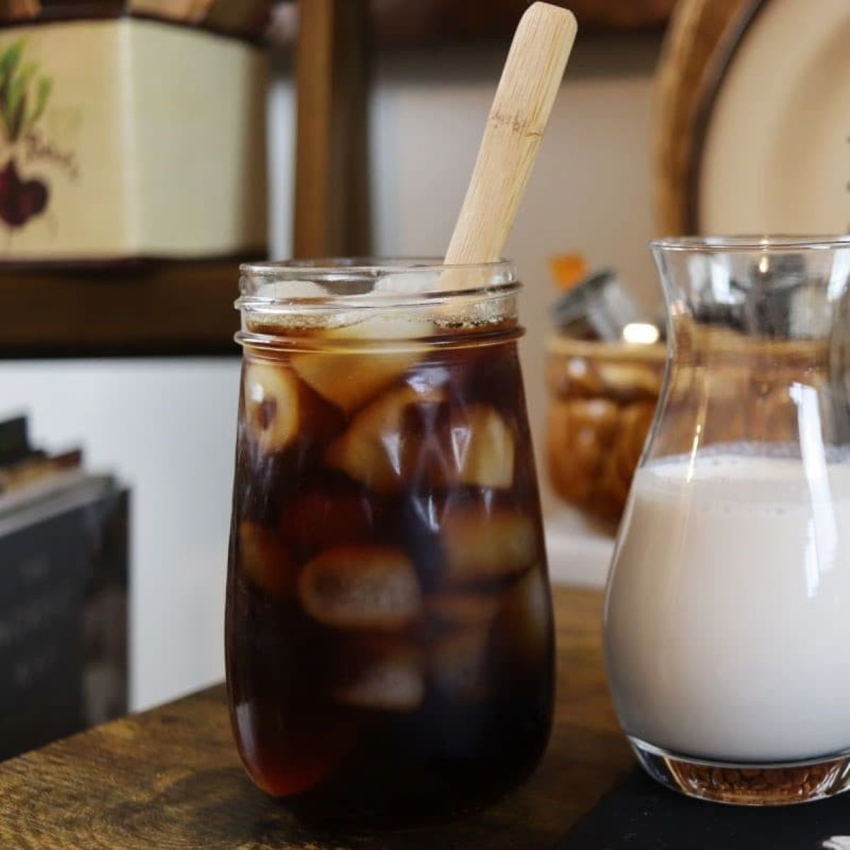 A mason jar filled with iced coffee and a wooden stirrer sits on a table next to a glass pitcher of milk. The background shows kitchen decor and containers.