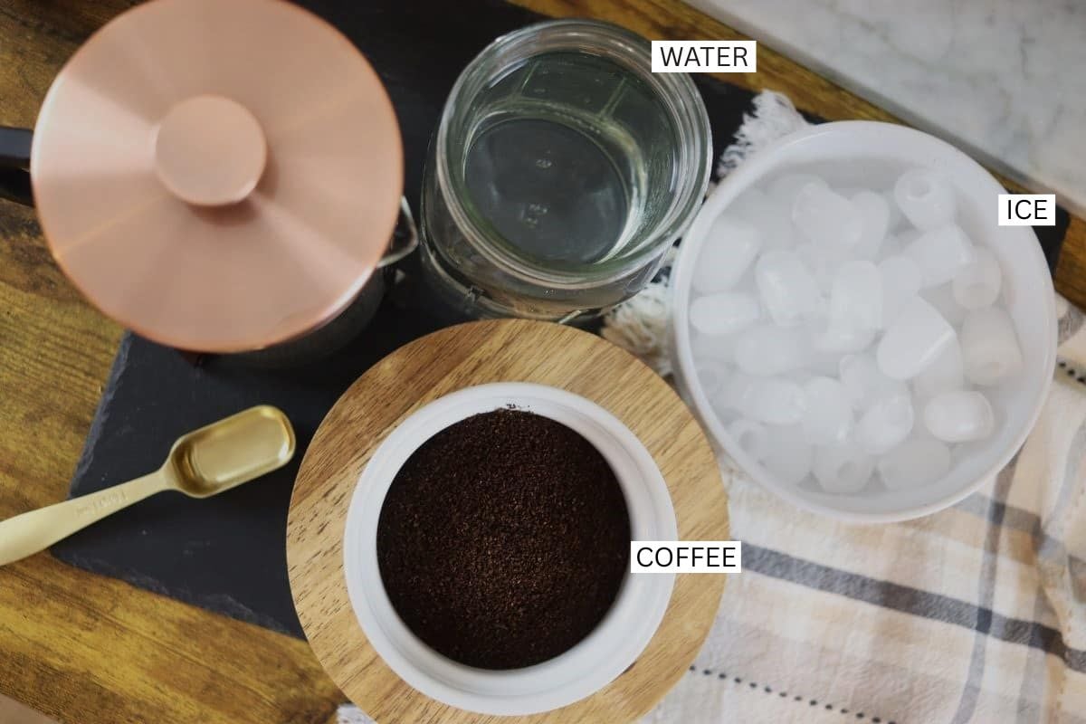 A labeled top view of a French press, a jar of water, a bowl of ice, a bowl of ground coffee, and a gold coffee scoop on a striped cloth. Labels identify Water, Ice, and Coffee.