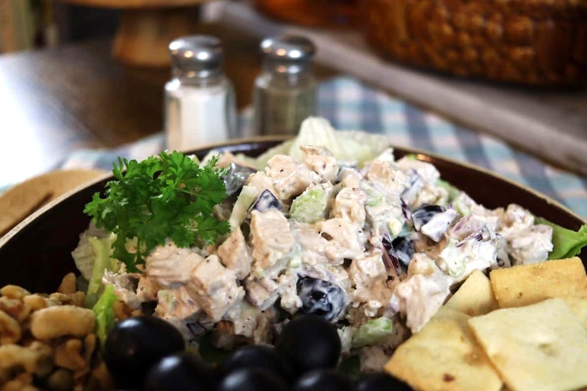 A close-up of a chicken salad with diced chicken, black grapes, celery, and a creamy dressing, garnished with parsley, and served with crackers and walnuts on the side. Salt and pepper shakers are blurred in the background.