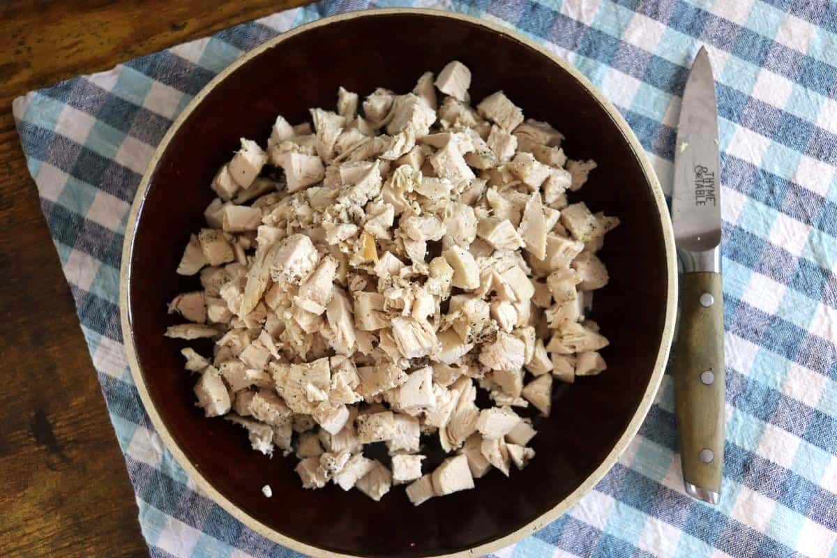 A brown bowl filled with chopped cooked chicken pieces sits on a blue and white checkered cloth; a knife with a wooden handle rests nearby on the cloth.