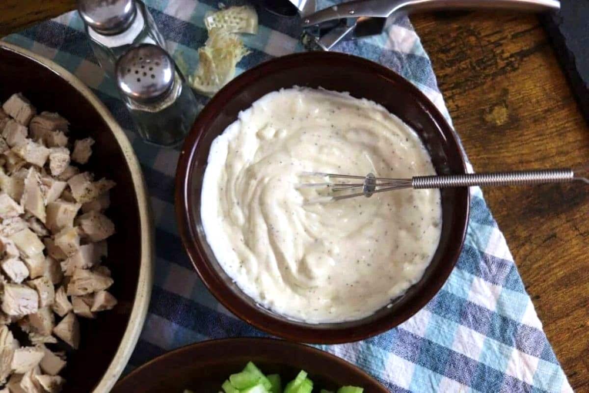 A small whisk rests in a bowl of creamy white dressing, surrounded by bowls of chopped chicken and celery on a blue plaid cloth, with salt and pepper shakers nearby.