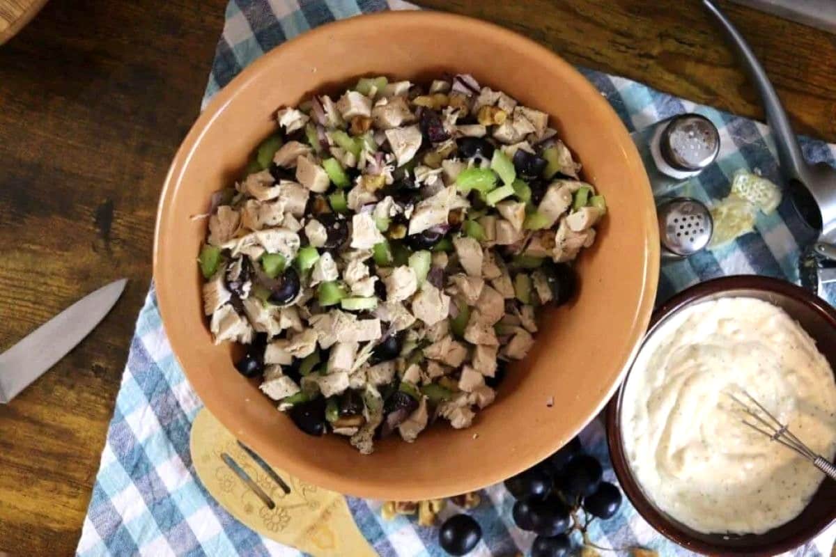 A bowl of chicken salad with celery and grapes sits on a checkered cloth next to a bowl of creamy dressing, grapes, walnuts, a knife, and salt and pepper shakers on a wooden table.