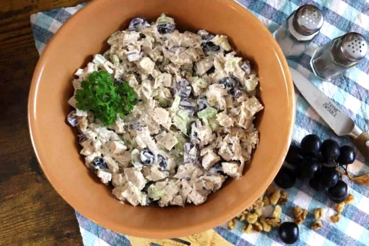 A bowl of chicken salad with grapes, celery, and walnuts, garnished with parsley, sits on a checkered cloth next to salt and pepper shakers, a knife, grapes, and walnuts.
