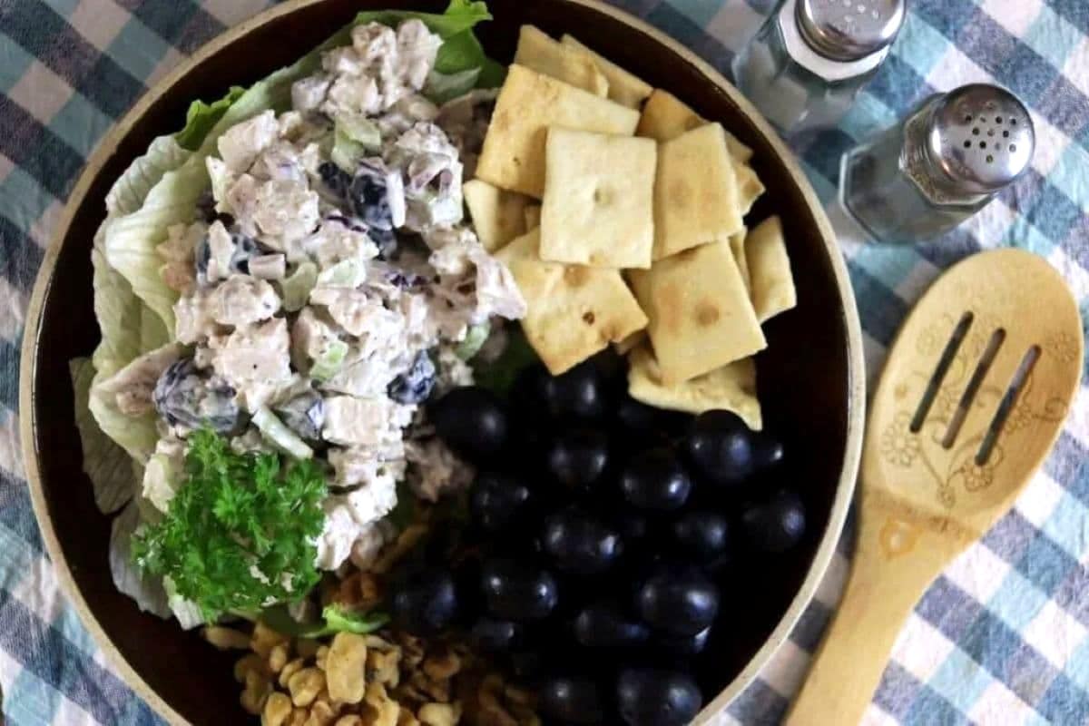 A bowl with chicken salad, black grapes, square crackers, lettuce, parsley, and nuts, placed on a checkered tablecloth next to salt and pepper shakers and a wooden spoon.