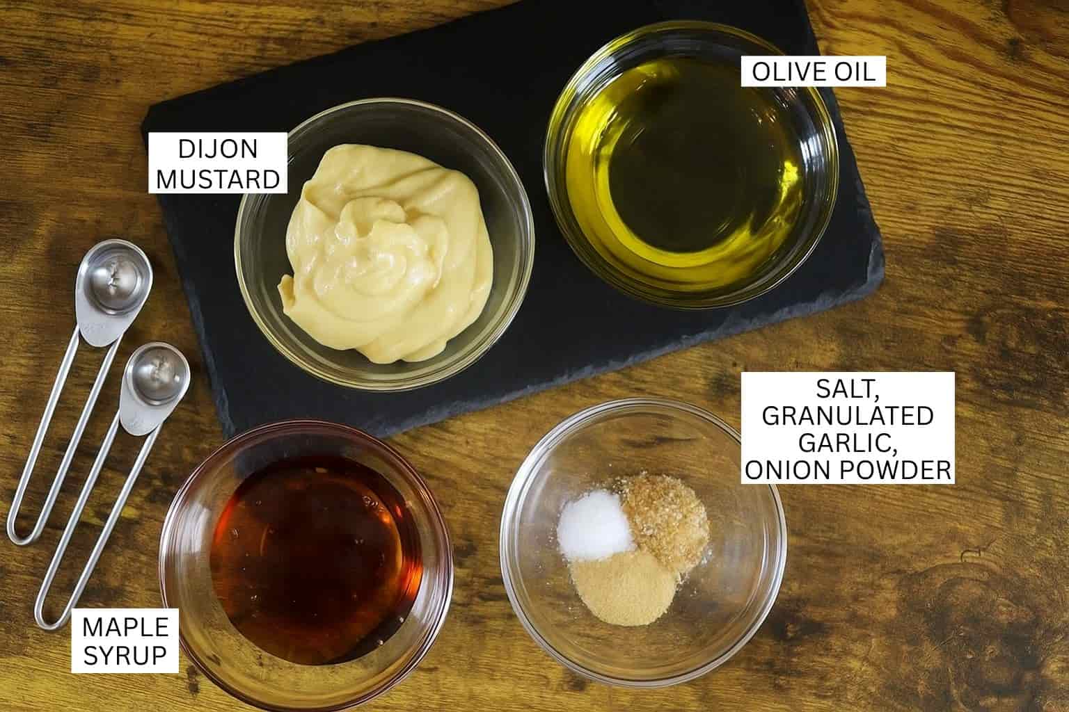 Four bowls on a wooden surface contain olive oil, Dijon mustard, maple syrup, and a mix of salt, granulated garlic, and onion powder; measuring spoons are next to the bowls.