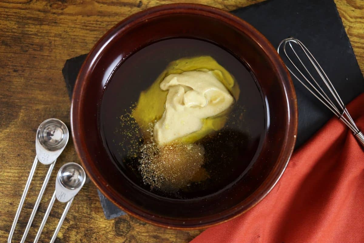 dijon mustard, olive oil, maple syrup and seasonings in a bowl sitting on a table next to measuring spoons and an orange cloth.
