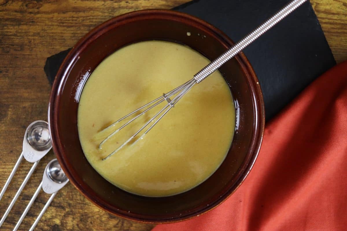 a bowl of yellow homemade dressing with a whisk in it sitting next to an orange cloth.