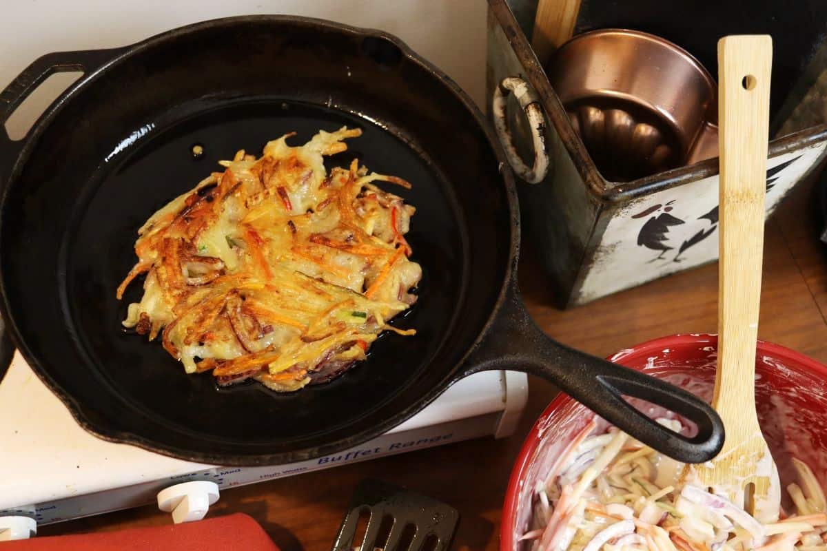 vegetable pancake cooking in a cast iron skillet on a stovetop with a bowl of julienned vegetables in batter and kitchen decor next to it.
