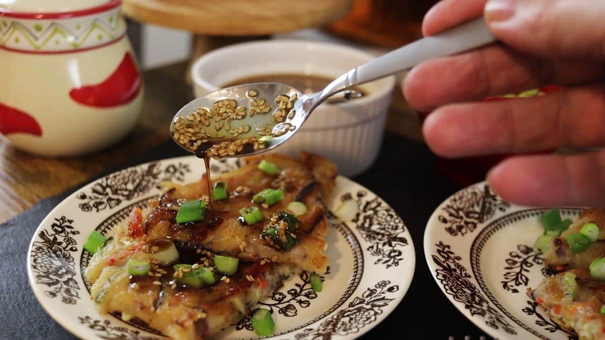 A hand drizzles sauce with sesame seeds over a slice of savory pancake topped with chopped green onions on a decorative plate, with a cup and dipping sauce bowl nearby.