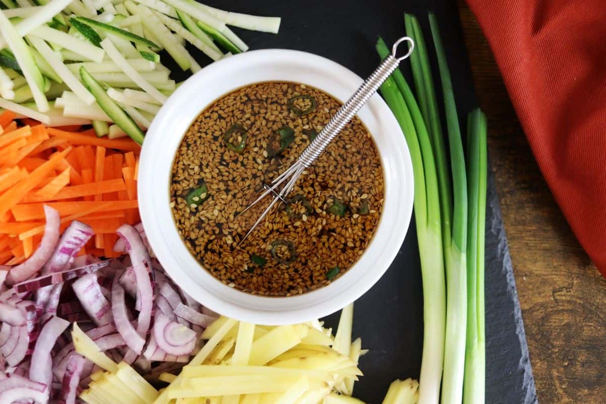 dipping sauce with sesame seeds and scallions in a bowl with a whisk in it sitting next to scallions and julienned zucchini, carrots, onions and potatoes.