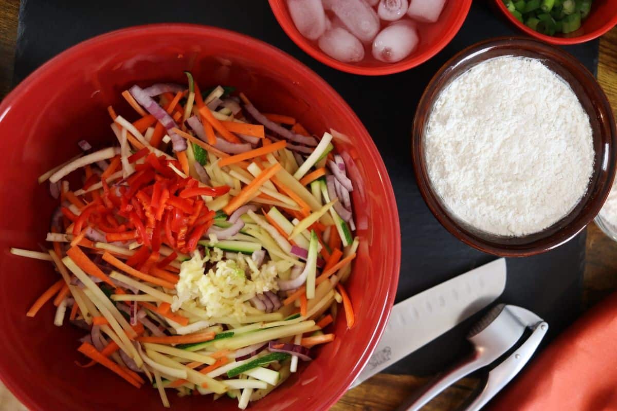 vegetables cut julienne style in a mixing bowl next to a bowl of ice water, a bowl of flour, a knife, and a garlic press.