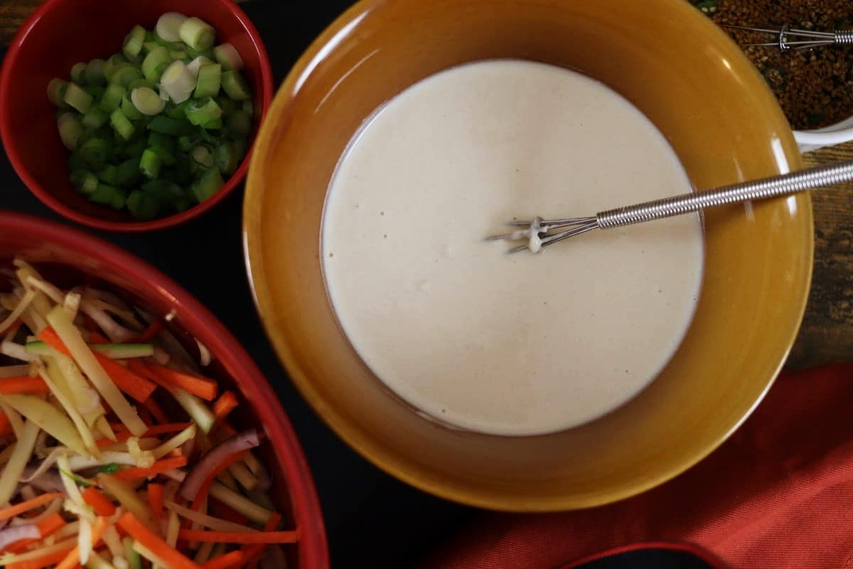 white batter in a bowl with a whisk next to a bowl of scallions and a bowl of julienned vegetables.