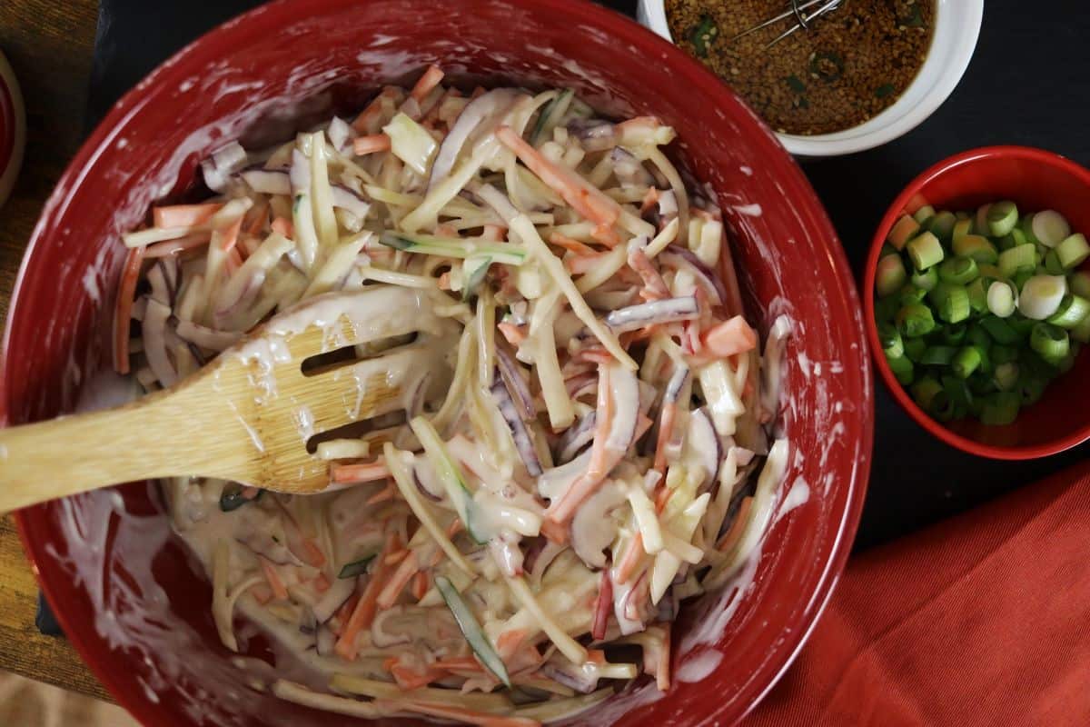julienned vegetables mixed with a batter in a bowl with a wooden spoon in it sitting next to a bowl of scallions and a bowl of sauce.