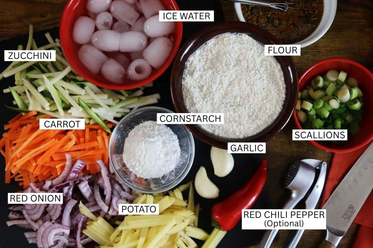 Various chopped vegetables (zucchini, carrot, red onion, potato, scallions, red chili pepper), garlic cloves, bowls of flour, cornstarch, and ice water are arranged on a wooden surface, each labeled.