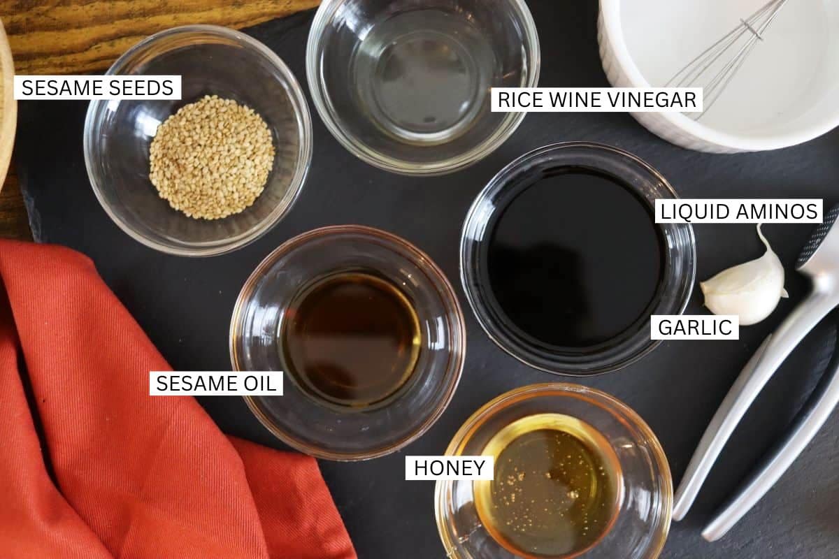 Five small bowls containing sesame seeds, rice wine vinegar, liquid aminos, sesame oil, and honey are arranged on a dark surface, with a garlic clove and fork nearby. Each ingredient is labeled.