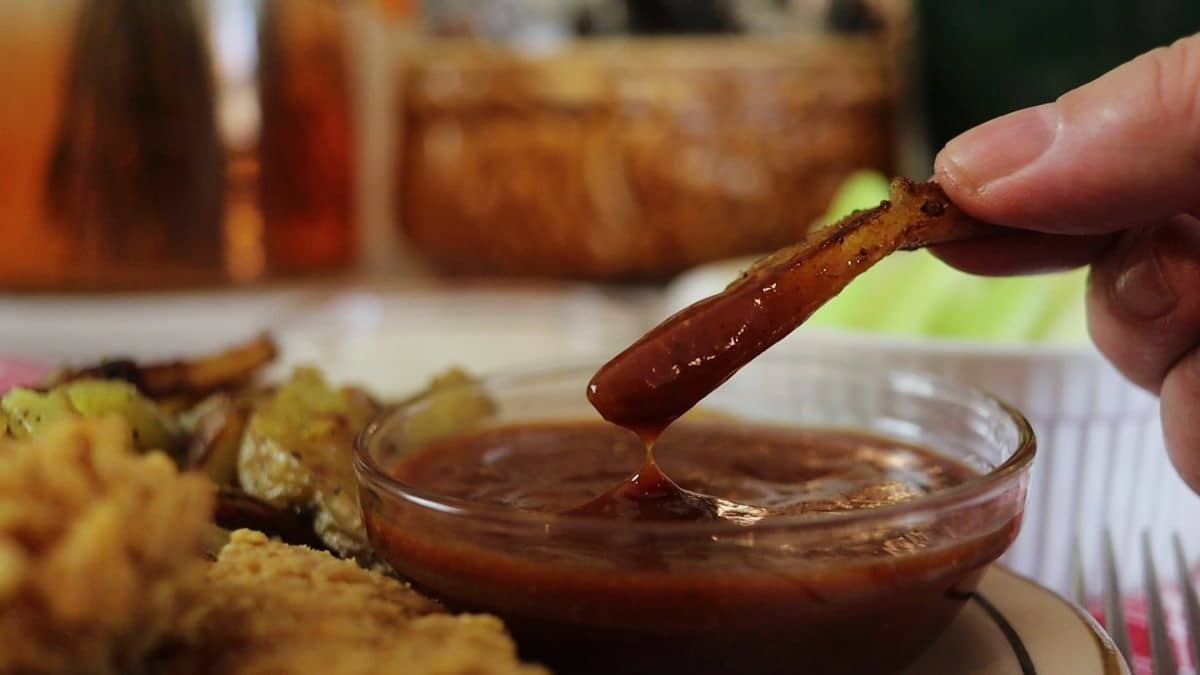 A hand dips a French fry into a small glass bowl of chicago mild sauce, with other fried foods and vegetables blurred in the background.