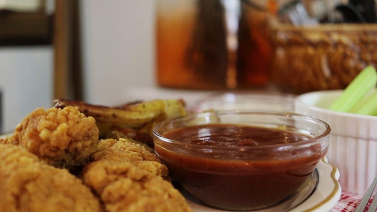 Small glass dish of dark reddish brown sauce with breaded chicken fingers and french fries visible next to it.