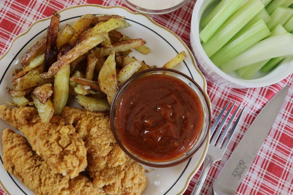 A plate with fried chicken tenders, seasoned fries, and a bowl of Chicago mild sauce, next to a bowl of celery sticks, on a red checkered tablecloth with a fork and knife beside the plate.