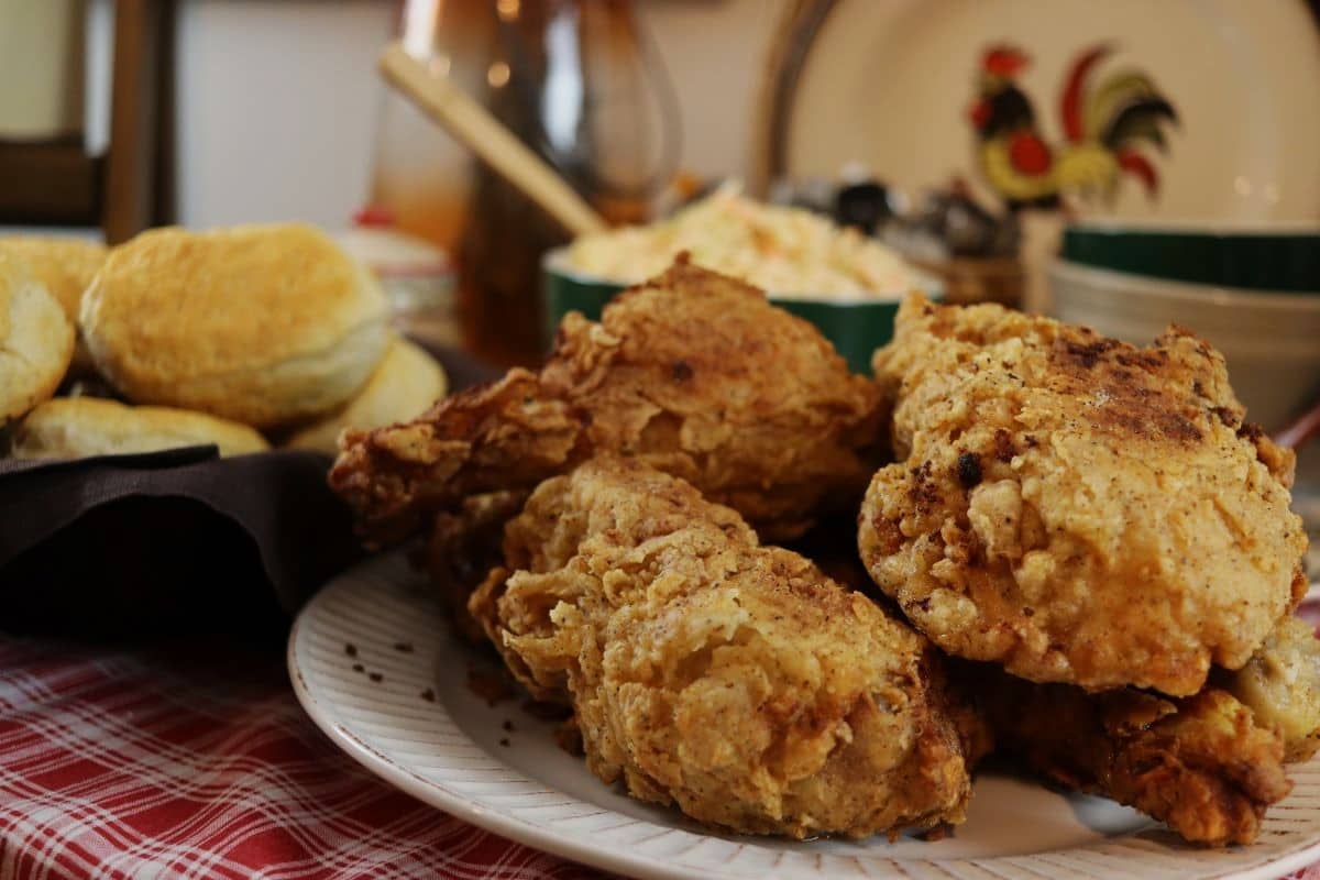 A plate of crispy fried chicken is in the foreground, with a basket of biscuits to the left and a bowl of coleslaw in the background on a red plaid tablecloth.