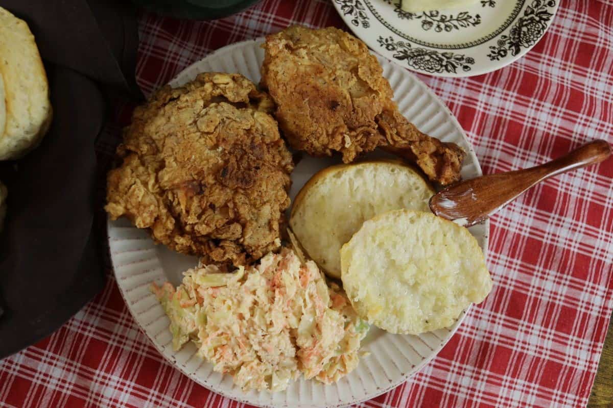 A plate with fried chicken, a split biscuit, and a serving of coleslaw sits on a red plaid tablecloth, with a wooden spreader resting on the biscuit.