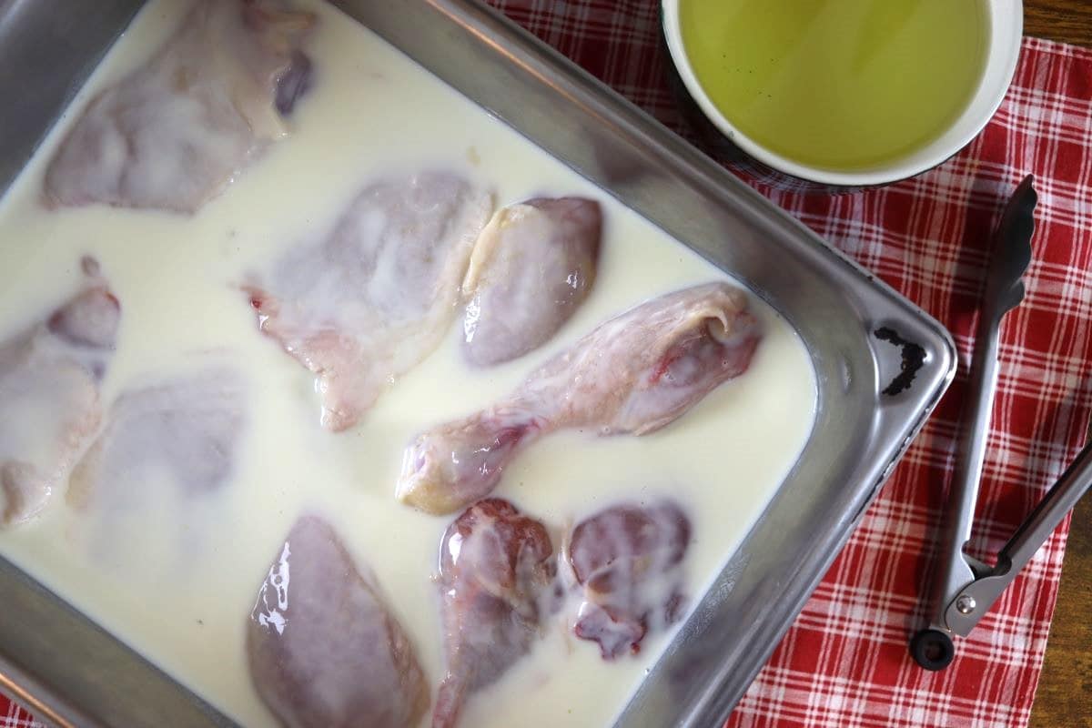 Raw chicken in a pan of buttermilk marinade sitting on a red checkerd cloth with a small bowl of oil visible to the side.