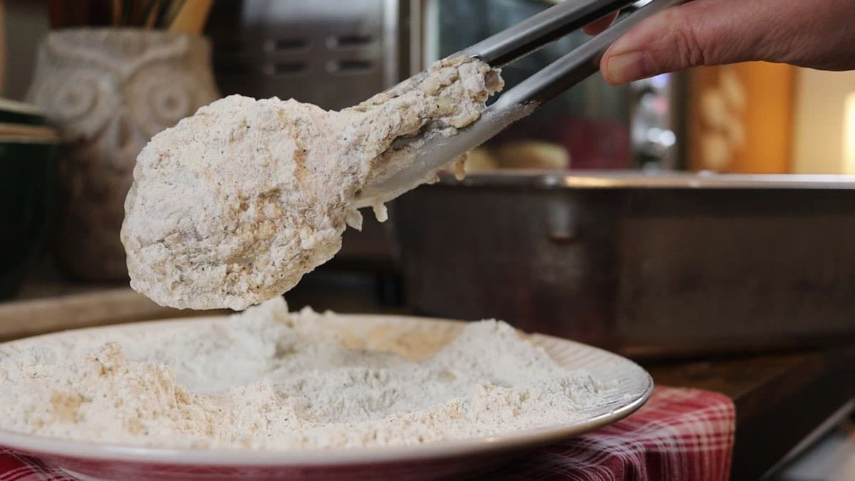 Tongs lifting a piece of chicken coated in flour mixture from a plate of the same flour mixture.