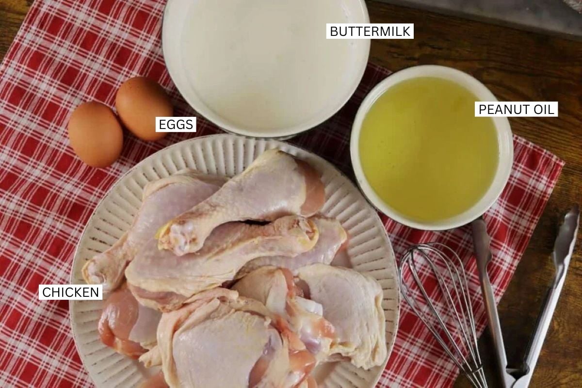 A plate of raw chicken pieces, two eggs, a bowl of buttermilk, and a bowl of peanut oil sit on a red plaid cloth, with labels identifying each ingredient. A metal whisk is beside the bowls.