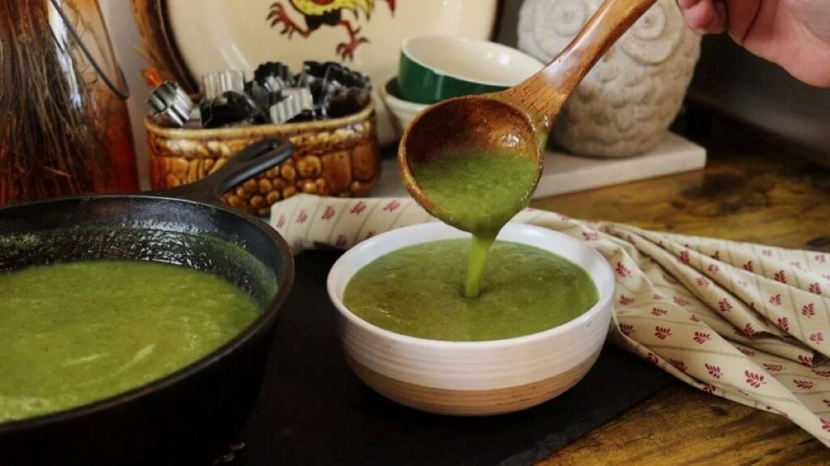 A person pours green soup from a wooden ladle into a white bowl, with a pot of soup and kitchen items in the background on a rustic wooden surface.