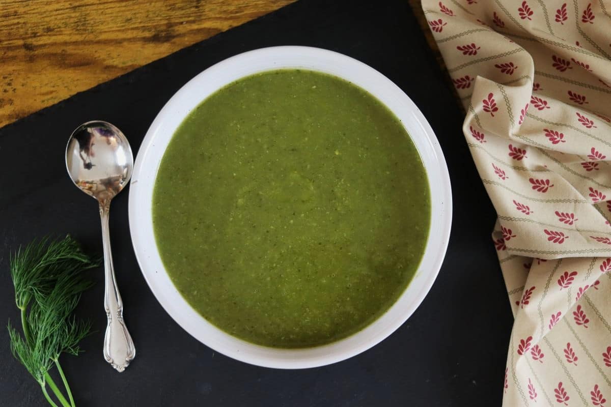 bowl of green broth sitting on a table with a soup spoon and a sprig of dill.