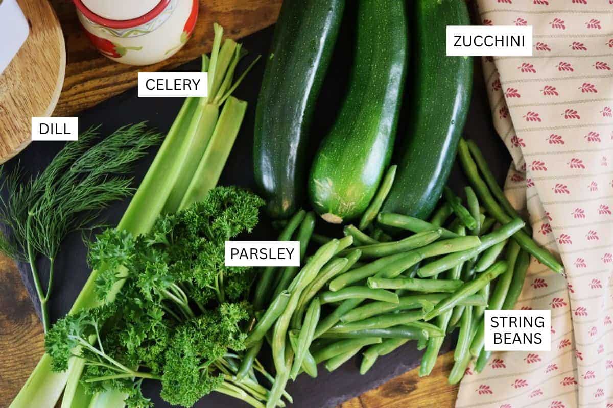A black tray with fresh zucchini, string beans, parsley, celery, and dill, each labeled with text. The vegetables are arranged on a wooden table beside a patterned cloth.
