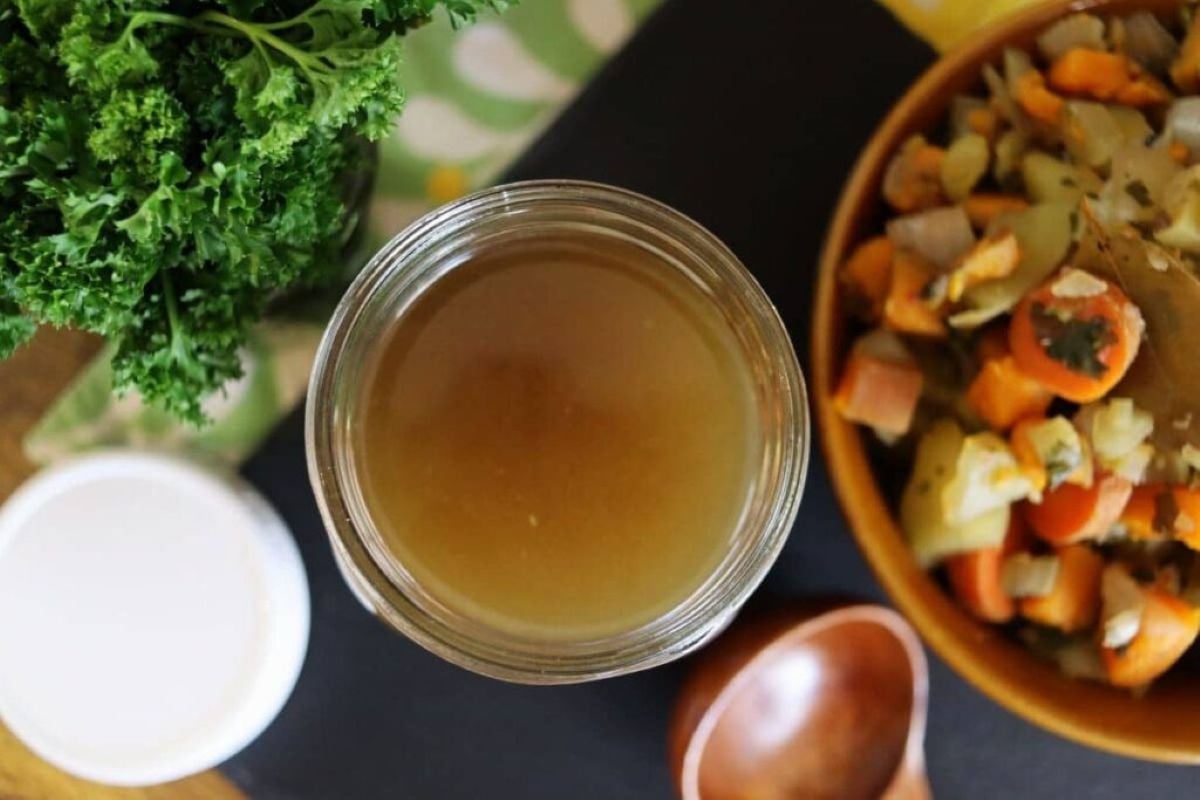 A jar of light brown vegetable broth sits on a dark surface next to a bowl of chopped vegetables, a bunch of fresh parsley, a wooden spoon, and a white lid.