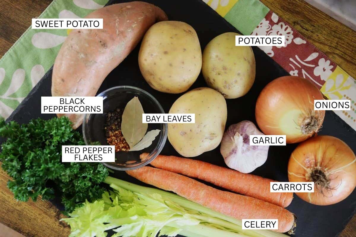 A group of vegetables and seasonings labeled on a black surface: sweet potato, potatoes, onions, garlic, carrots, celery, parsley, black peppercorns, red pepper flakes, and bay leaves.