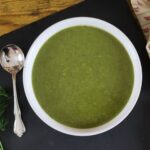 Bowl of green broth on a slate board with a soup spoon next to it.