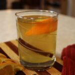 Glass with yellow liquid, an orange wedge, and a cinnamon stick in it sitting on a striped wooden cutting board.