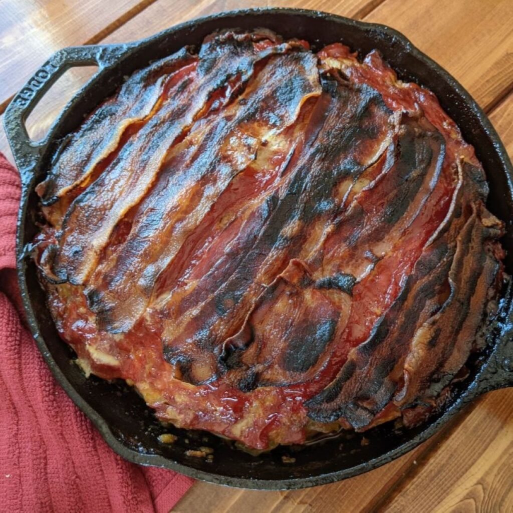 A cast iron skillet filled with meatloaf, topped with overlapping strips of crispy, browned bacon, sits on a wooden table next to a red cloth.