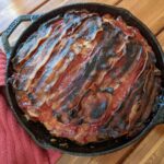 A cast iron skillet filled with meatloaf, topped with overlapping strips of crispy, browned bacon, sits on a wooden table next to a red cloth.