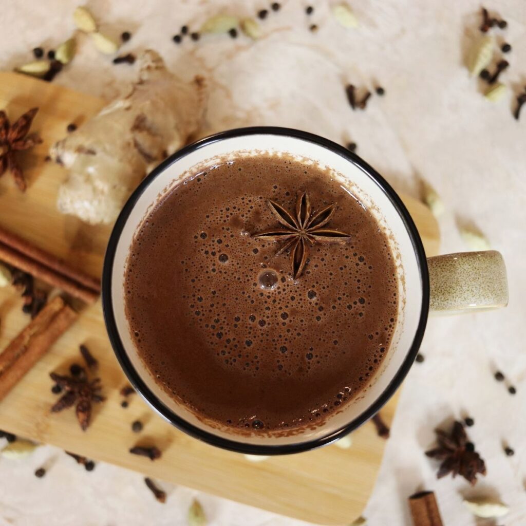 Mug full of brown liquid with an anise star floating in it and anise stars, cinnamon sticks, and cloves scattered on the counter around it.
