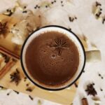Mug full of brown liquid with an anise star floating in it and anise stars, cinnamon sticks, and cloves scattered on the counter around it.